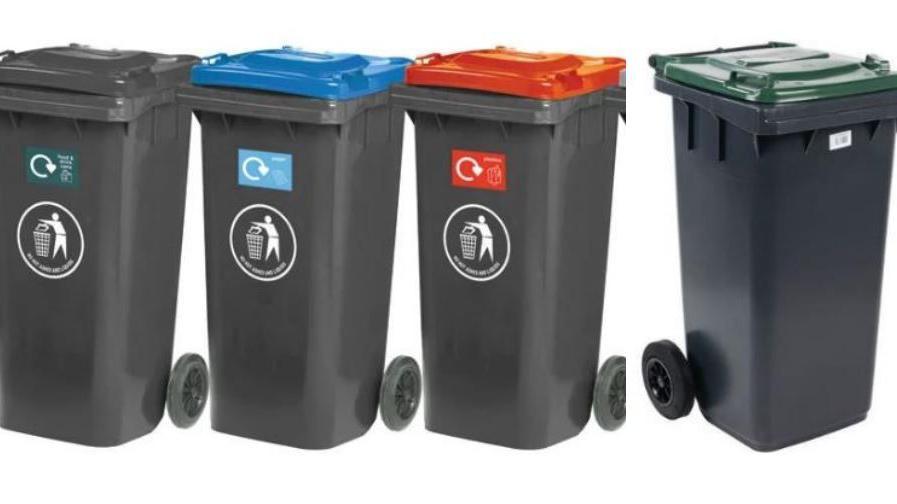 Four black wheelie bins in a row. The bins all have grey bodies, but each have different coloured lids. Left to right the colour of the bin lids are grey, blue, red and green