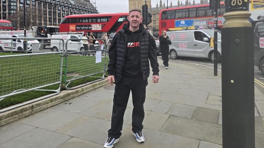 Gary Quinn, a painter and decorator, stands on pavement in central London. with traders driving in vans behind him at a rally. Three red London buses are also behind him.