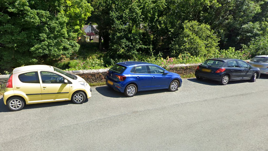 A street view of Market Road in Tavistock. There is a river wall next to which are parked several cars, and trees behind the wall