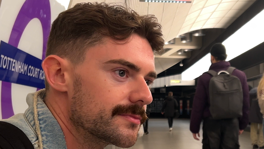 Dan Harry, a young man with a moustache and wearing a denim jacket, is pictured sitting on a bench at Tottenham Court Road Tube station. He gazes off camera and is facing the tracks. Other people walk on by