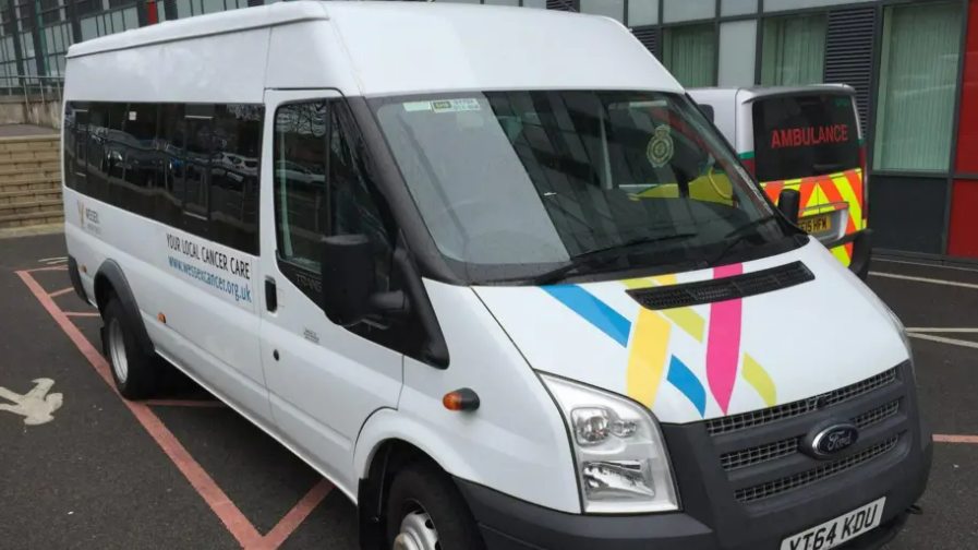 A white Ford Transit minibus with Wessex Cancer Support branding parked outside a hospital, with an ambulance beside it.