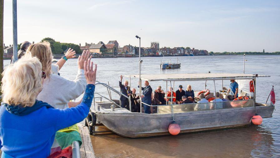 The West Lynn ferry with people aboard. There are also people at the dock waving them off.