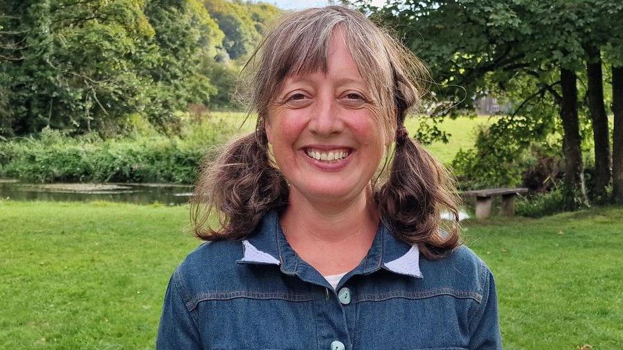 A smiling Lorna Rees standing near the river. She has her hair in bunches and is wearing a denim shirt