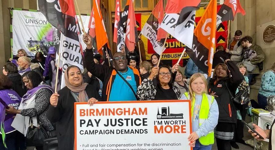 A group of about a dozen people, all holding trade union flags, which are orange and black or red and white. A group of four people in the centre of the image holds a sign that says Birmingham Pay Justice Campaign Demands.