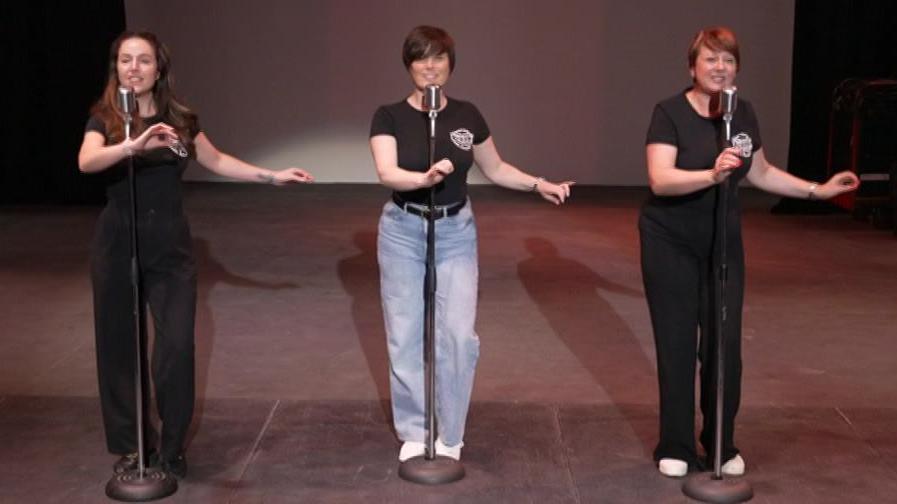 The Boogaloo Babes during rehearsals. The three women each stand behind a microphone stand while singing and dancing, their left arms outstretched.