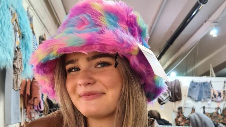 A smiling Amelia Butlin standing in a clothing shop posing at the camera in a multi-coloured fur hat with the price tag attached.