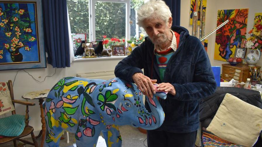 Sutton has white hair and a grey short beard. He is looking towards the camera with his hands resting on the trunk of an elephant statue. He is wearing a dark navy fleece jacket with a patterned jumper beneath and a light coloured shirt colour visible. The statue is painted a bright blie and has colourful yello and green flowers painted all over it. He is standing in a room with a large window behind him through which dark trees are visible and the walls of the room are decorated with several brightly coloured works of art and paintings.