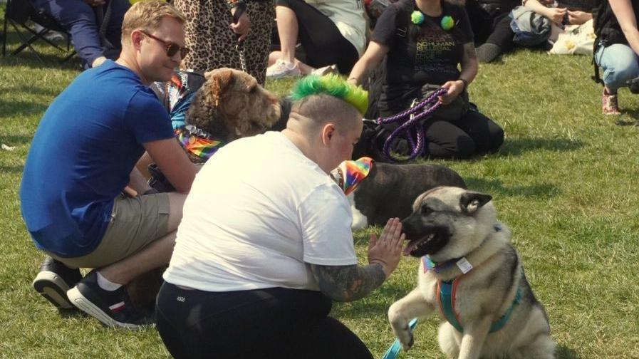 Pride festival-goers with their dogs in the gardens in Leamington, showing one dog in a rainbow collar with their foot up to their owner