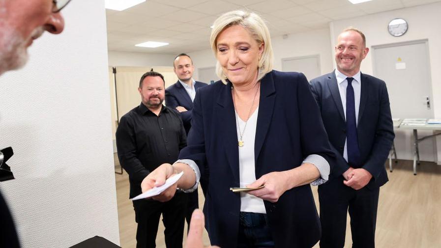 President of the 'Rassemblement National' (RN) group at the National Assembly Marine Le Pen (C), flanked by Steeve Briois (R), mayor of Henin Beaumont, delivers her identity papers to vote for the European Parliament election at a polling station in Henin-Beaumont, north of France