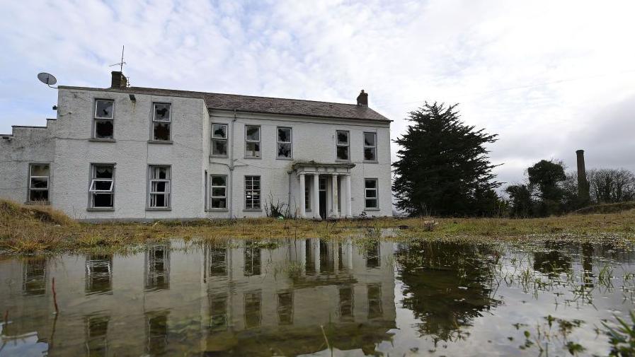 The former Marianvale Mother and Baby home in Newry. A dilapidated two-storey building surrounded by overgrown grounds and a holy cross plinth.