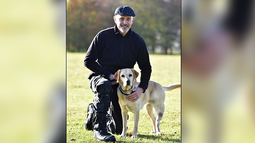 Ian Partington, wearing all black, including a black cap, kneeling on one leg, holding a blonde coloured dog. He is in a park.