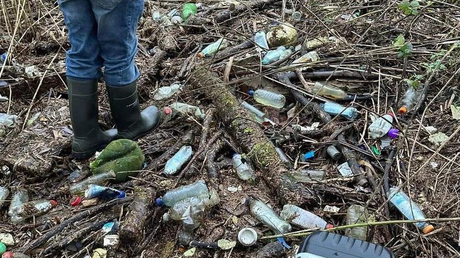 Riverside scene showing dozens of plastic bottles and tin cans left as rubbish, while a person stands in wellie boots 