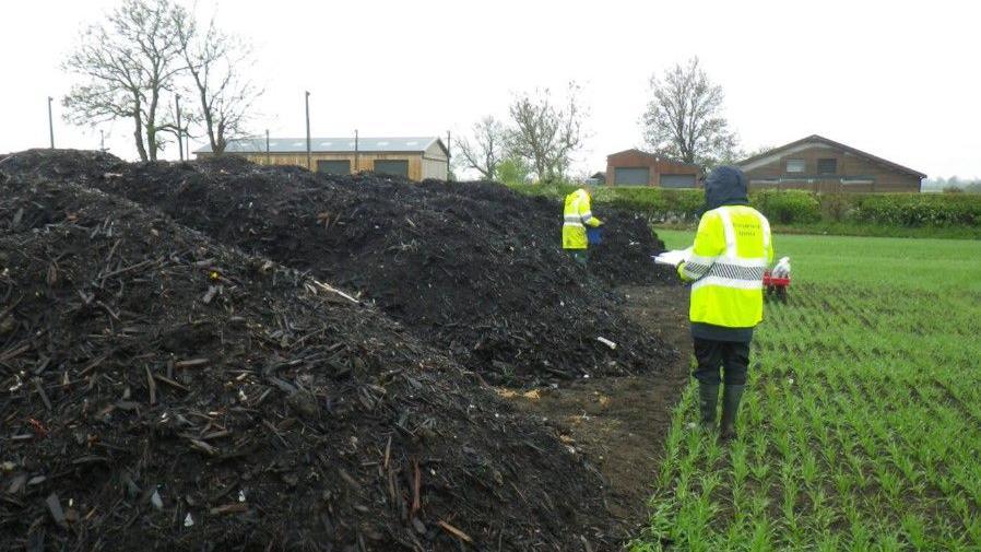 Environment Agency officers in high-vis jackets standing in a field next to a pile of compost.