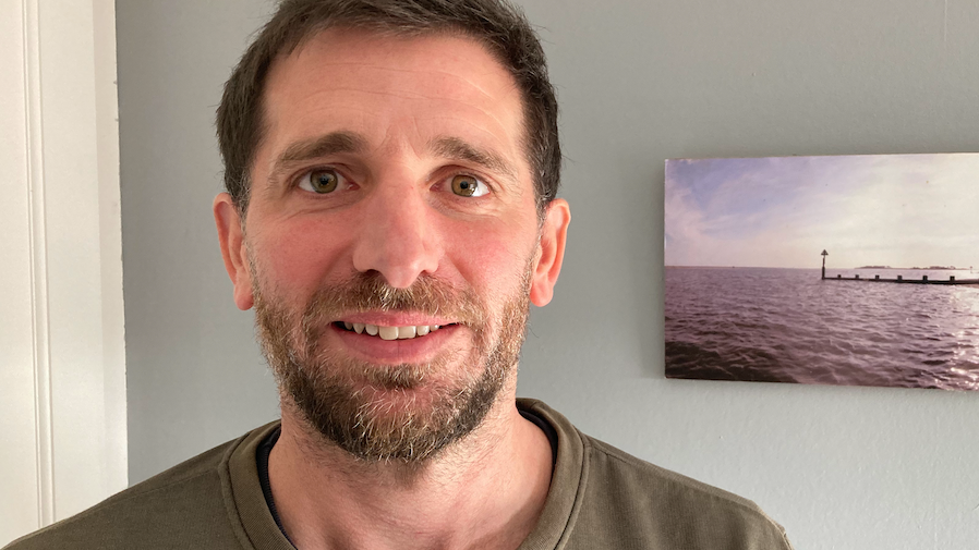 Head and shoulders of a man with short brown hair, and a short brown beard, standing in front of a plain backdrop, with half a slim painting of an ocean scene behind him. 