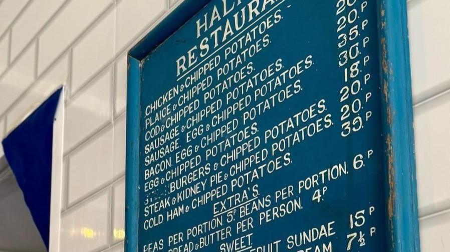Blue wooden framed board hangs on a white tiled wall with the prices of items on sale at a fish and chip shop