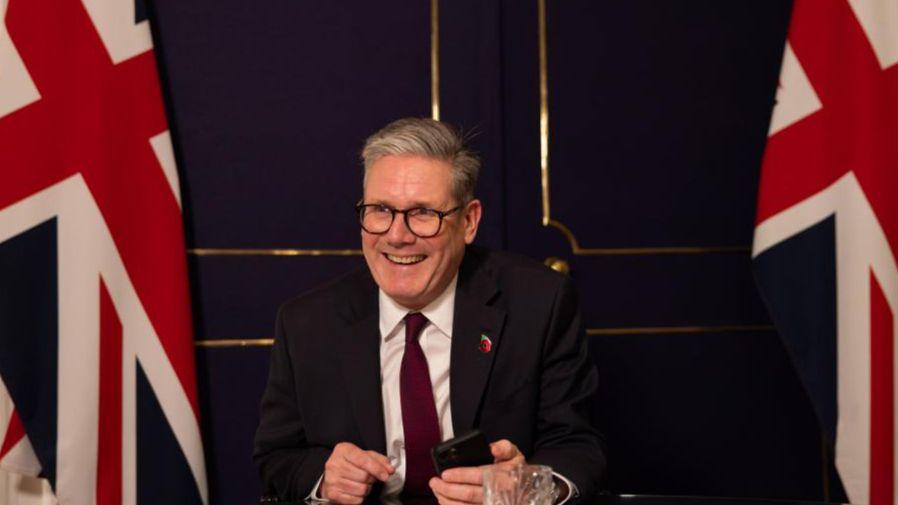 The prime minister speaking to Donald Trump from Downing Street in November 2024, following his US election victory. He is wearing a dark suit, white shirt and red tie and smiling as he sits at a table, holding a mobile phone in his left hand. Two UK flags can be seen in the background