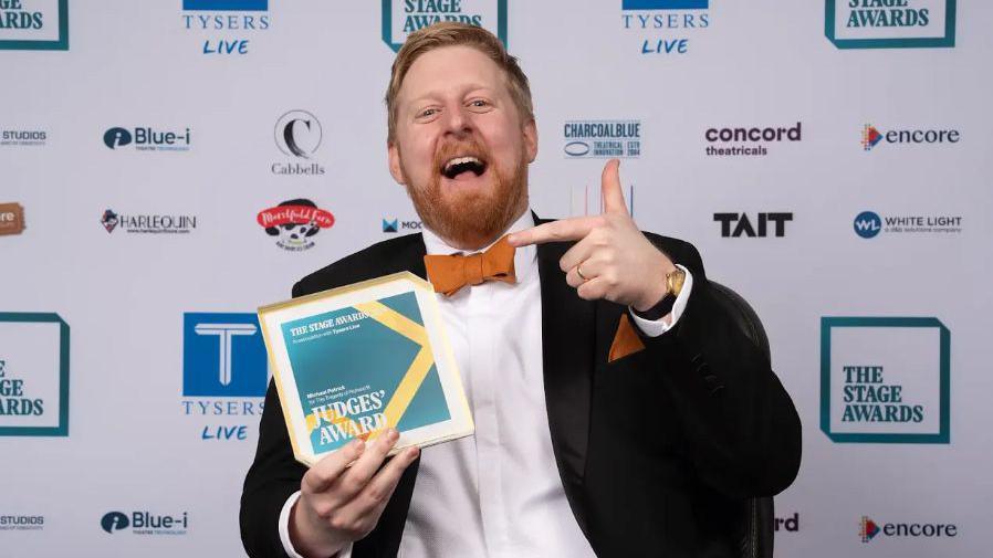 Michael Campbell is wearing a black suit, white shirt and orange bow tie. 
He is seen holding the Judges' Award from the Stage Theatre Awards.