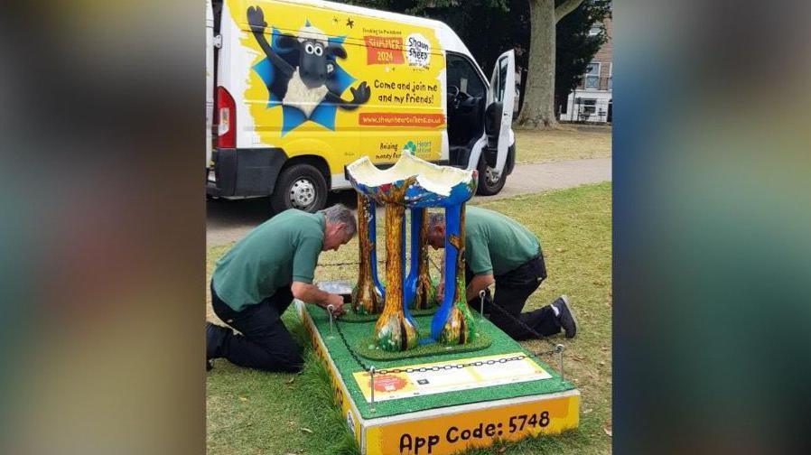 Two men in green shirts tend to a colourful statue which has been smashed up and only the legs of a sheep remain. 