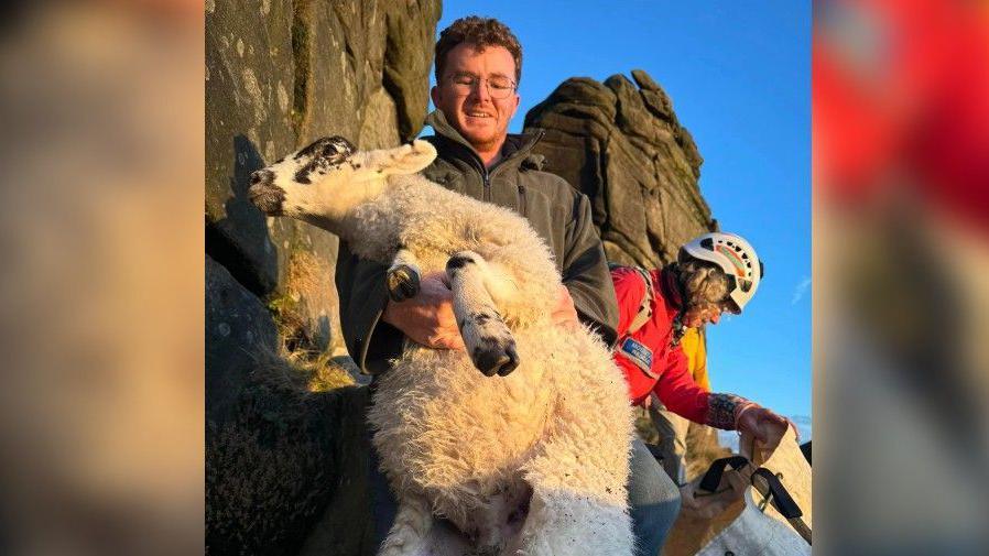 A man with glasses carrying a sheep down a cliff face alongside a mountain rescue member dressed in red