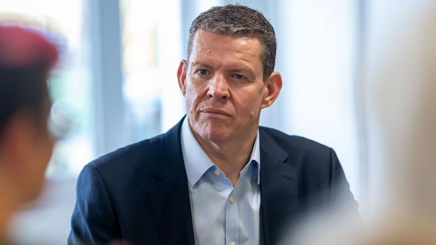 Plaid Cymru leader Rhun ap Iorwerth in conversation with an unknown person, who is out of focus in the foreground of the picture. He is wearing a dark blue suit with a light blue shirt with the top button undone and no tie