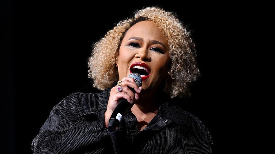 Emeli Sande has blonde, curly hair and is wearing a black top. She is singing into a mic. There is a black background.