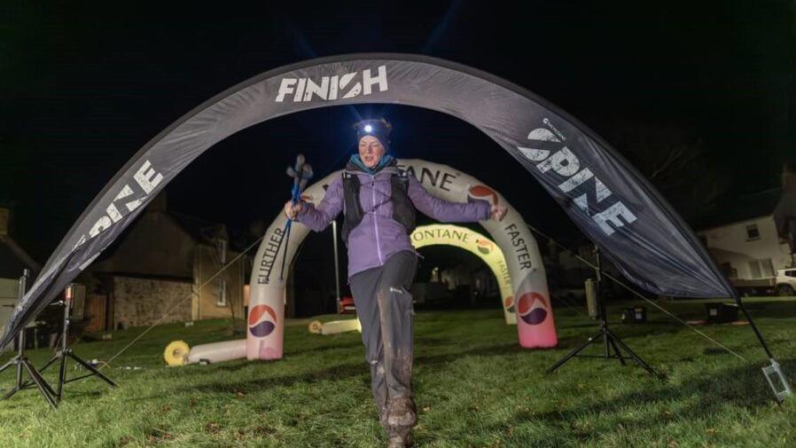 A runner crosses a race finishing line in the dark. She is wearing mountain-climbing gear, including muddy tracksuit bottoms and a helmet with a light on.