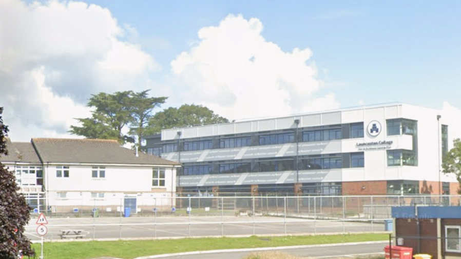 Mid shot image of Launceston College building on a sunny day. An outdoor area for students is located in front of the building and is enclosed by metal barriers. A small road in front of the building and runs alongside a small patch of green grass. 