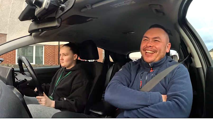 Andrea, in a black hoodie and hair pulled in a bun, sits in the driver's seat of the instruction car beside instructor Nicky Lui, a man with short dark hair, laughing as he gives instructions on starting the car. He wears a blue fleece. The interior of the car is light grey.
