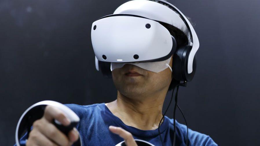 A man uses the PlayStation VR 2. It is larger than the other two headsets, and it is a sleek white curved shape.