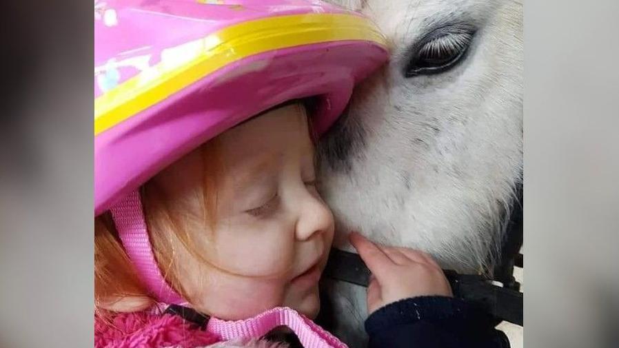 An up close image of Aoife, wearing a pink helmet, cuddling the face of a white Shetland pony