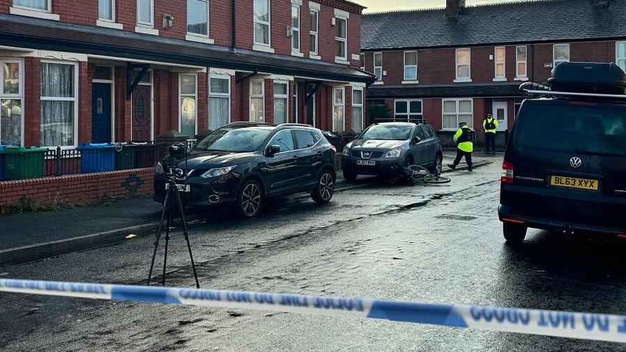 Police cordon strapped across street with two police officers and a tripod seen near parked cars outside terraced houses.
