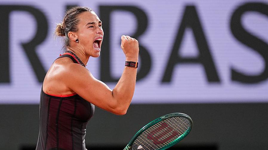 Aryna Sabalenka, wearing a black dress and holding her green racquet in her left hand, screams and clenches her right fist in celebration