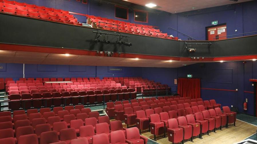 Rows of red theatre seats in the closed position set out in tiers over two levels.  Its walls are painted purple.