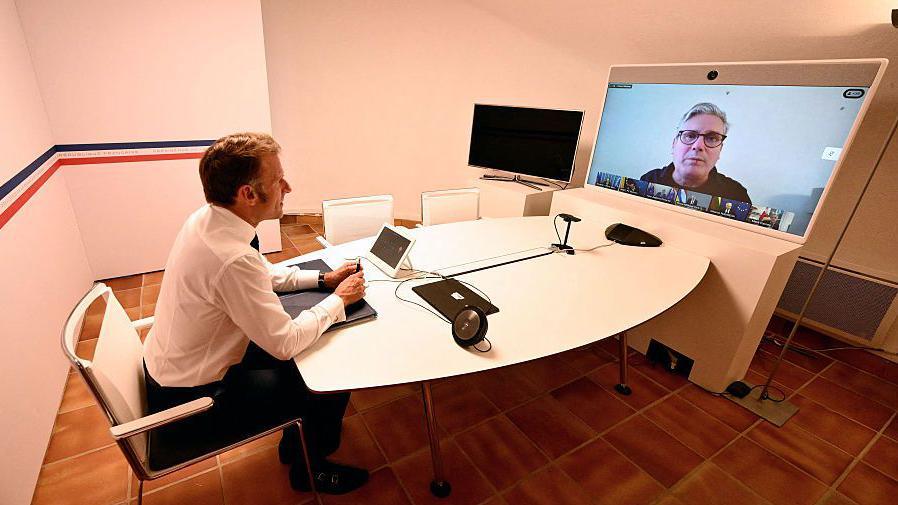 Emmanuel Macron sits at a table in front of a video conferencing screen. Macron has short brown hair and wears a white shirt and dark suit trousers. He is sat at a round white table in a white-walled room with brown tiled flooring. At the other end of the table is a large screen on which a video conference is being held. Sir Keir Starmer is enlarged on the screen, while a row of small boxes containing the other participants sits below him. Starmer wears a grey top and black-rimmed glasses, and sits in front of a plain white background.
