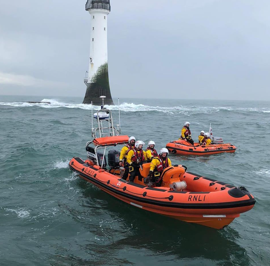 Two small lifeboats with crew sit out at sea, with a lighthouse in the background