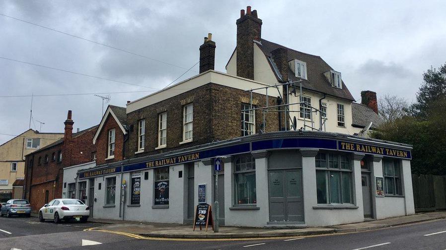 The Railway Tavern, which is a pub on the corner of a residential road. It is grey with a blue banner that runs along it. There are grey doors and windows.