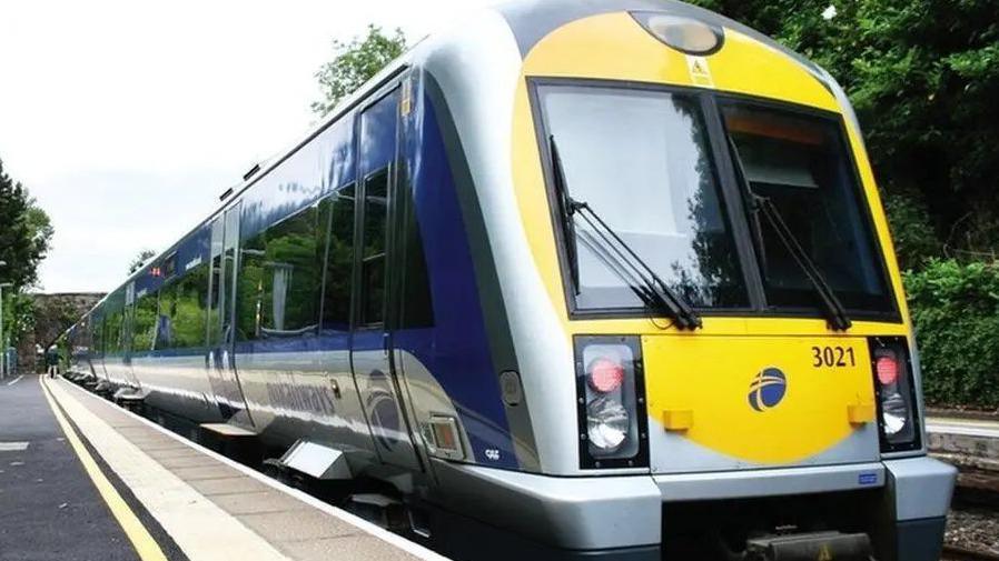 A Translink train stopped at a station in Belfast. The train is blue, yellow and grey and is pictured from the front at a slight angle. 
