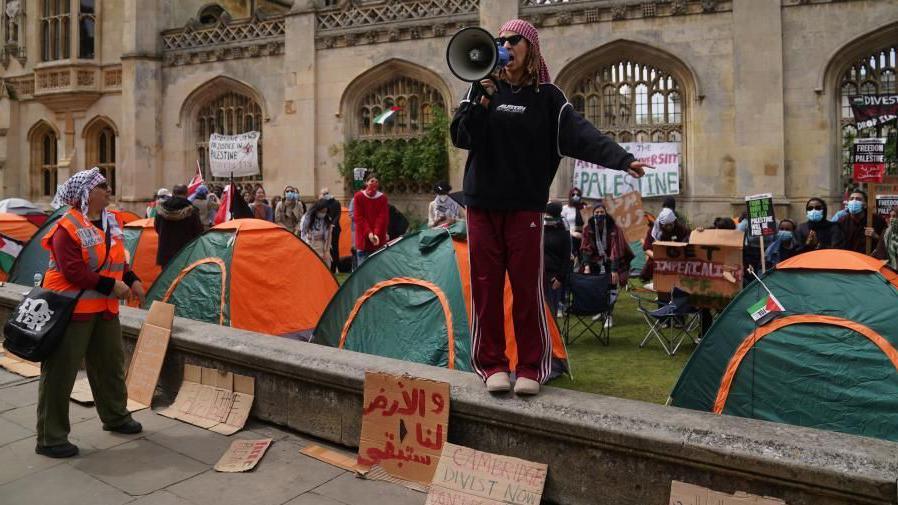A woman wearing a black top and red trousers stands on a wall in front of a collection of tents. She is holding a megaphone.