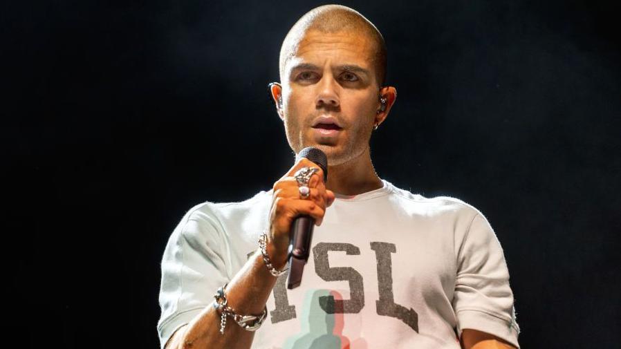 Max George singing into a microphone. He has a shaven had and earpieces in. He is wearing a short-sleeved t-shirt that says YSL on it, and he has silver bracelets on his right arm and silver rings on his right hand. He is stood on a stage with a black backdrop, looking past the camera at the audience.