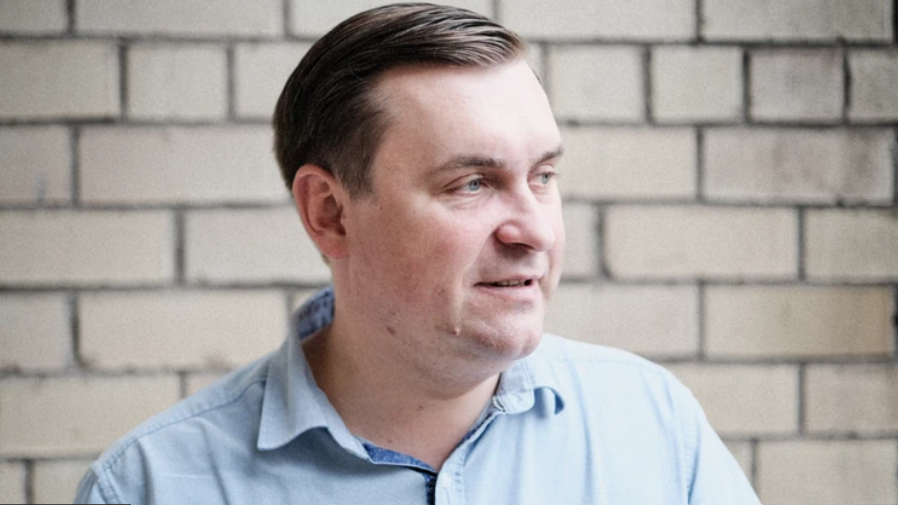A man in a blue shirt and dark hair stands in front of a white brick wall.