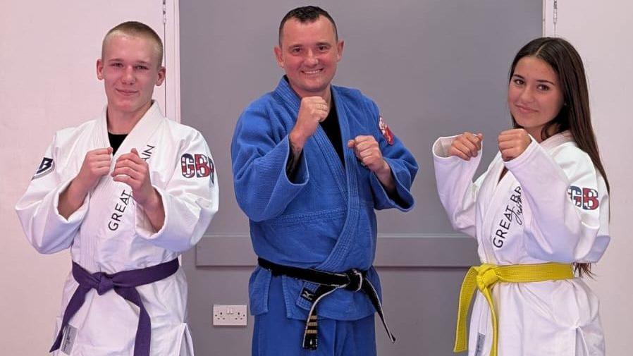 A teenage boy and a teenage girl are pictured in jiu-jitsu clothing with green and purple belts. They are standing either side of their sensei, who is wearing a dark blue uniform.