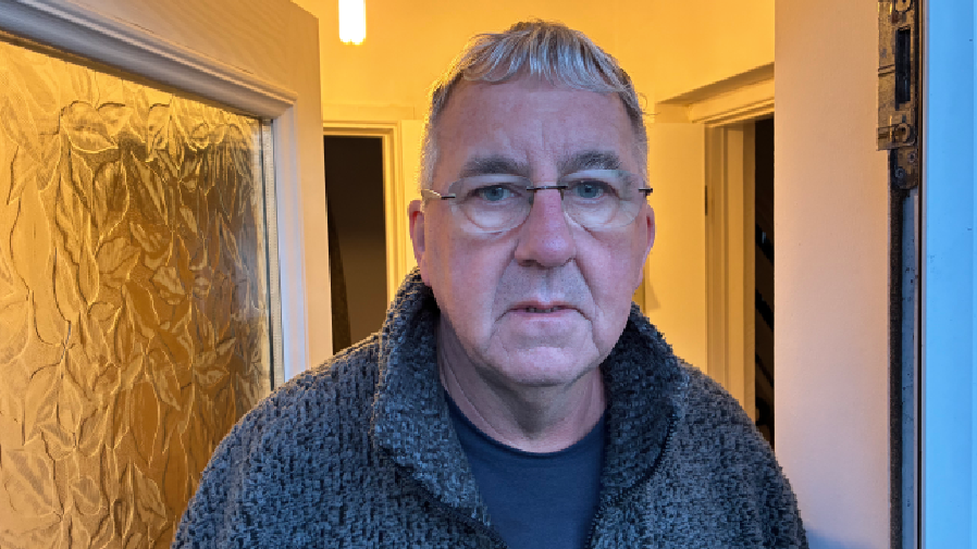 David Barlow - dyn 65 oed gyda gwallt brith byr a sbectol, yn sefyll o flaen drws blaen ei gartref