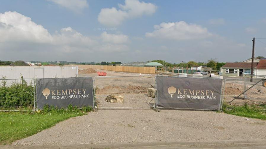 Construction work under way at the Kempsey Eco Business Park site in Kempsey. Two large signs can be seen at the front entrance. Gates and wooden fencing surround the circumference of the building site. Other buildings can be seen in the background.