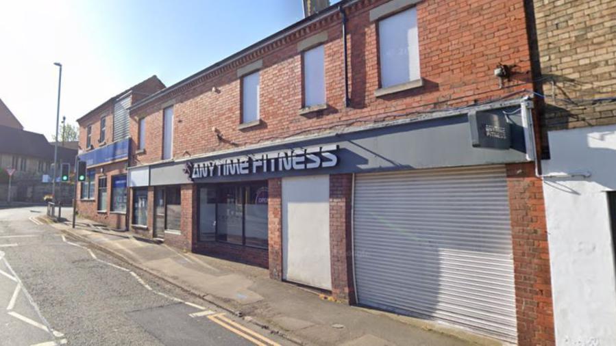 A long brick terrace with "Anytime Fitness" written above a doorway. A shutter is down on the shop front next to it.