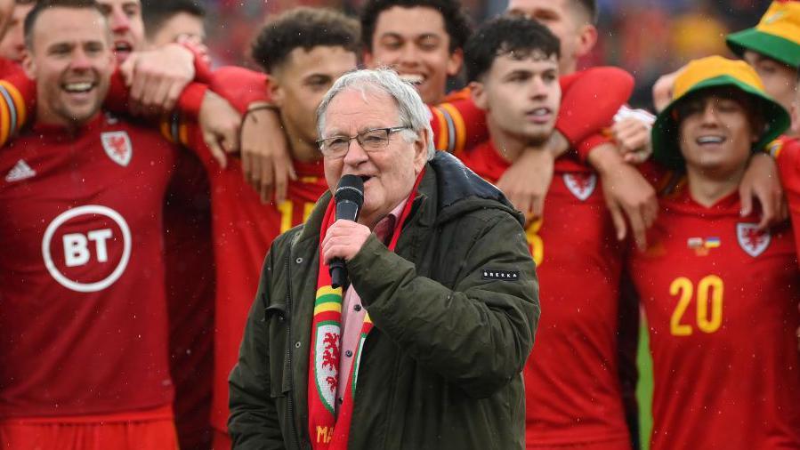 Dafydd Iwan singing passionately into a microphone on the pitch at a Wales game in Cardiff City Stadium. Behind him, a group of Welsh football players stand in a line side by side.