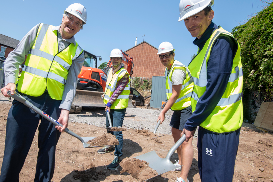 Four men wearing hard hats and reflective safety jackets hold spades full of soil