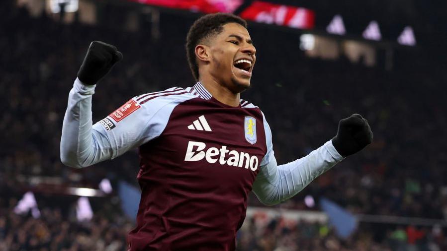 Marcus Rashford is wearing a claret and blue Aston Villa shirt as he holds up his arms and smiles