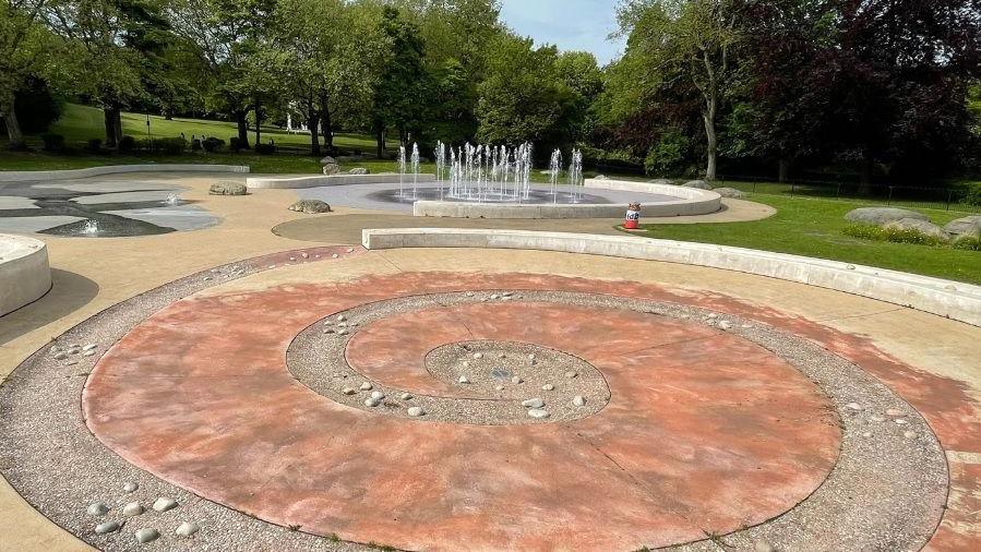 Spiral-shaped pool (drained of water) at Clifton Park splash pool in Rotherham, with pebbles in the spiral. Fountain beyond.