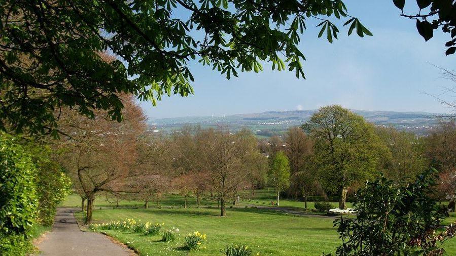 A path leads down the hill surrounded by greenery, bushes, trees and lined with daffodils. The path leads towards a playing field in Great Harwood. You can see hills and houses and wind turbines in the distance. 