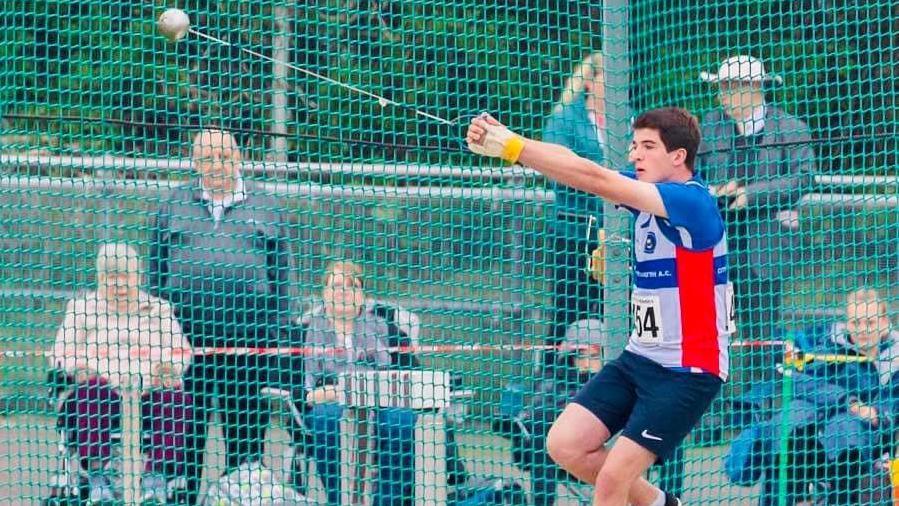 Jack Burgess mid-throw in-front of a crowd of spectators. He is standing within a green net and wearing a blue and white shirt with red stripes on the side